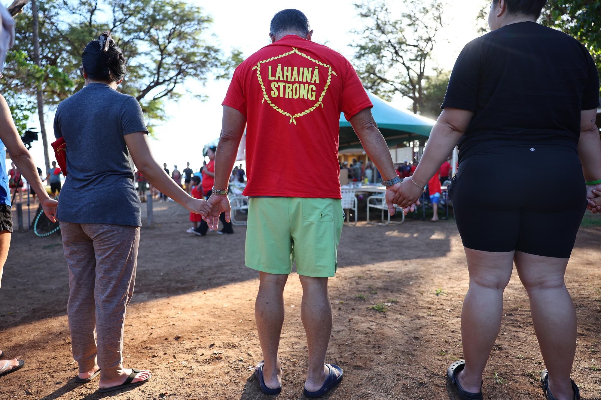 Community members hold hands in a prayer circle and a man wears a "Lahaina strong" T-shirt.