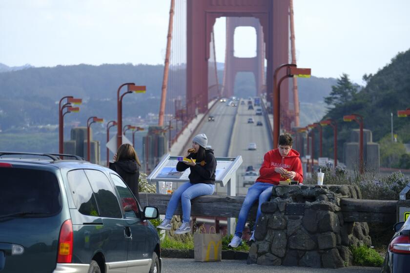 People eat while sitting at a vista point by the Golden Gate Bridge Friday, March 27, 2020, in Sausalito, Calif. The surge of coronavirus cases in California that health officials have warned was coming has arrived and will worsen, Gov. Gavin Newsom said Friday, while the mayor of Los Angeles warned that by early next week his city could see the kind of crush that has crippled New York.(AP Photo/Eric Risberg)