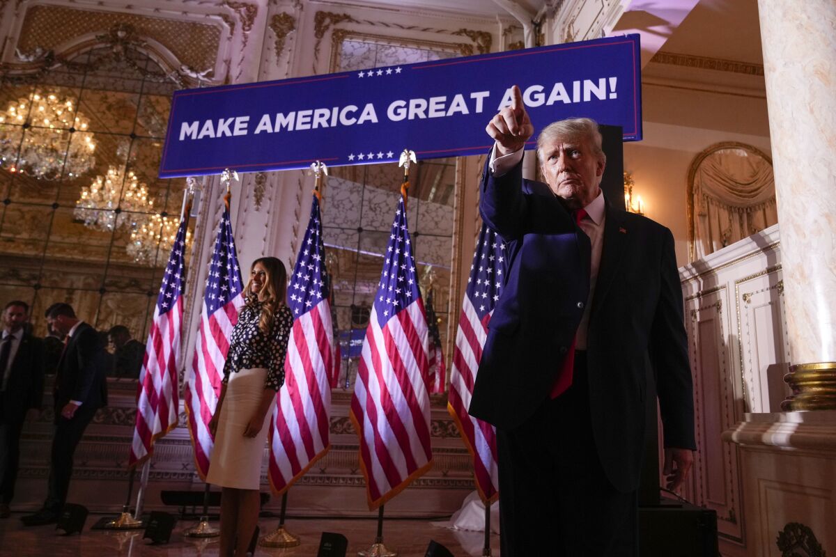 Donald Trump bir dizi bayrağın ve karısının önünde duruyor ve işaret ediyor 