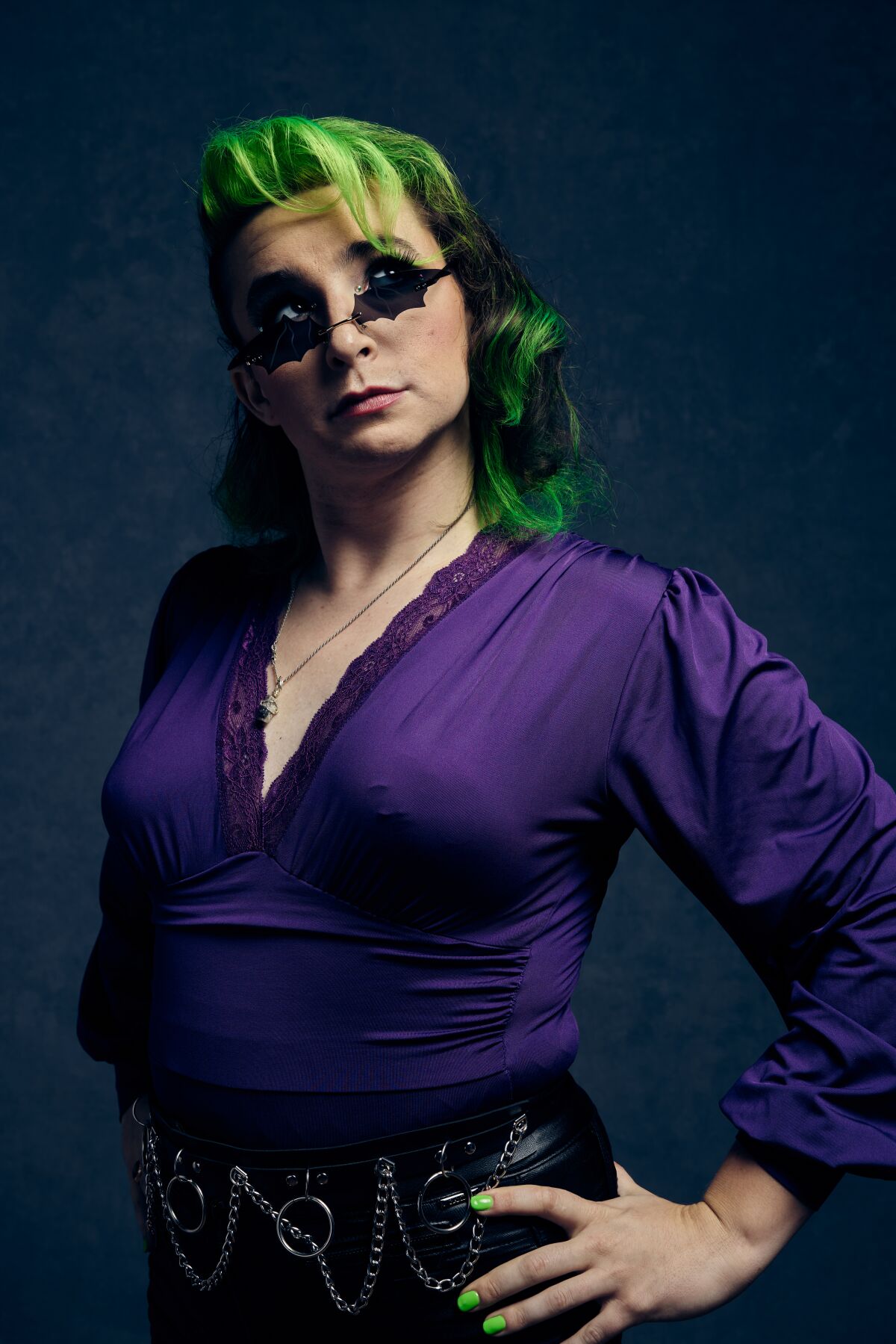 a woman in a purple silk top and green hair puts her hands on her hips and poses