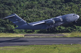 A gray military plane takes off from a tarmac, with greenery in the background