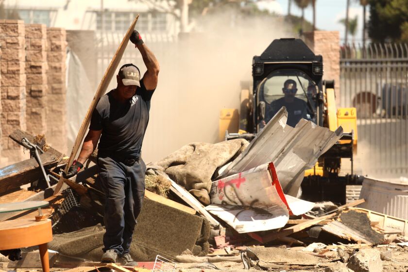 WATS, CA - AUGUST 12, 2022 - - Darren Burkhart, left, and Brian Car, in skid-steer loader, firefighters with the Los Angeles Fire Department Heavy Equipment unit, raze a homeless structure in a vacant lot in Watts on August 12, 2022. The homeless have been living in the vacant lot for years. After giving them notice days in advance the homeless who were living there left the encampments and some, who wanted housing, were given temporary housing by members of Homeless Outreach Program Integrated Care System (HOPICS). The vacant field that was meant to revitalize a community bled of its economic base and traumatized by the 1992 riots. It has been sitting without any development since '92. (Genaro Molina / Los Angeles Times)