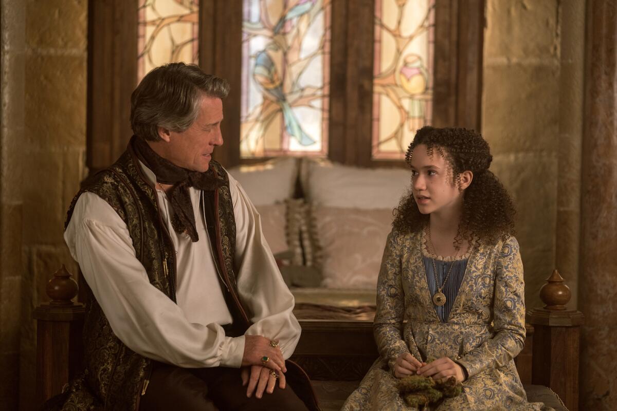 Wearing vaguely medieval clothing, a man with graying temples sits in conversation with a girl.
