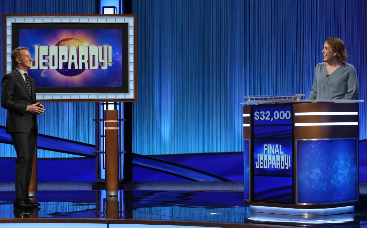 A man in a suit speaking to a woman standing behind a podium on the set of "Jeopardy!"