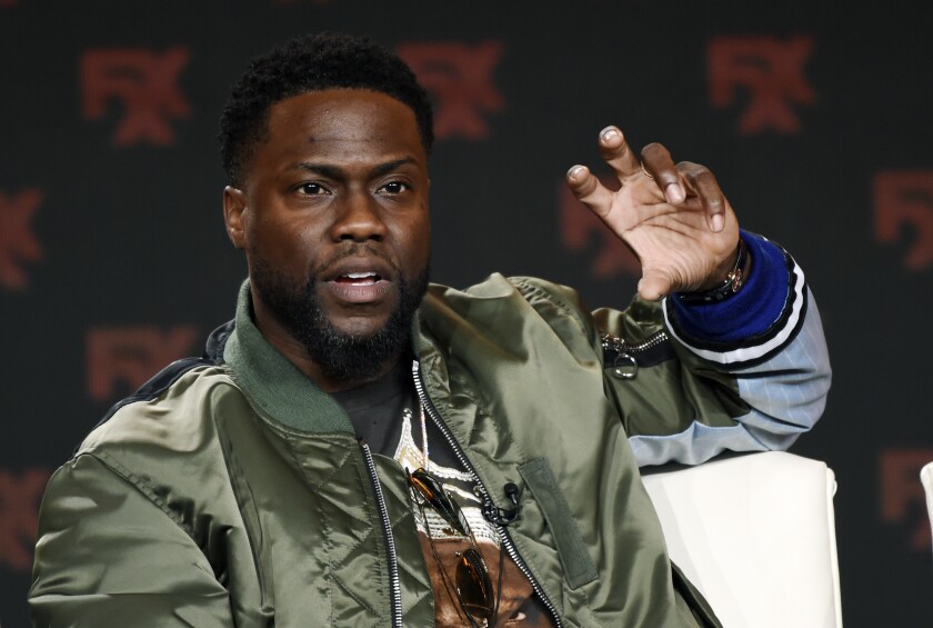 Kevin Hart in an olive-drab bomber jacket raising his hand