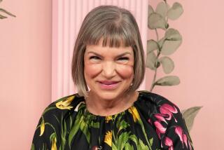 Mindy Cohn smiling in a black floral dress in front of a pink backdrop