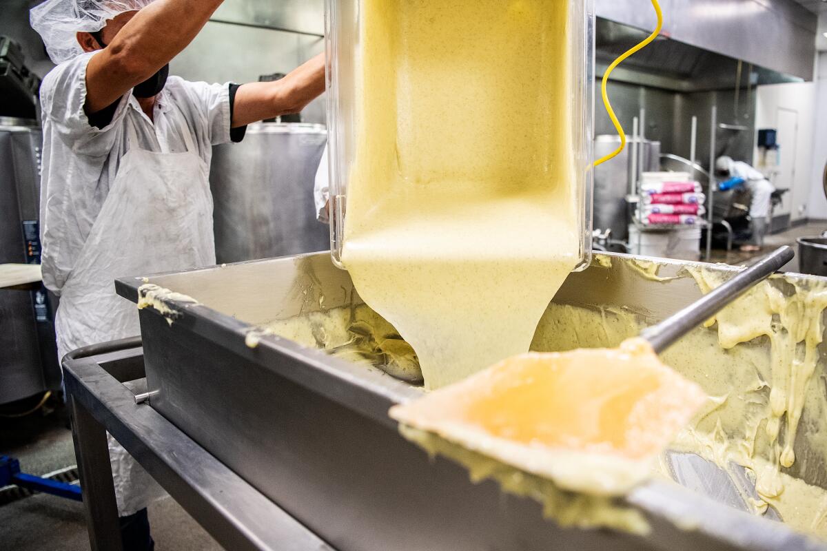 A worker in an apron pours a liquid from a plastic tub into a stainless steel vat.