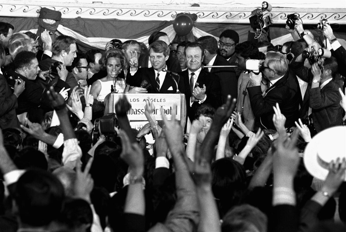 Robert Kennedy stands at a lectern behind a microphone, surrounded by people and media.