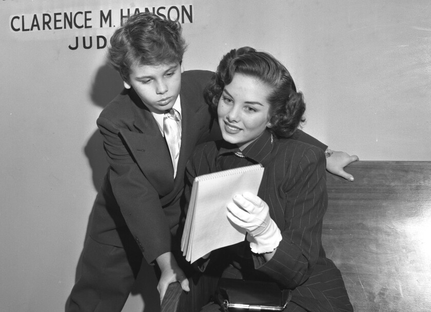 A boy in suit and tie leans over a young woman in a skirt suit and gloves. They look at a document she's holding.