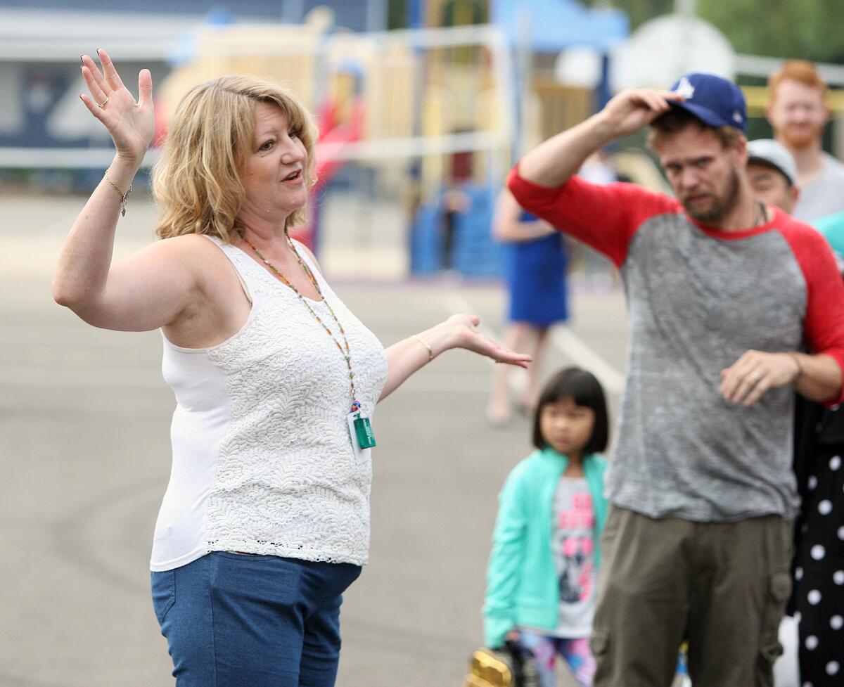 A woman in a white tank top speaks and gestures with both arms on a school playground.