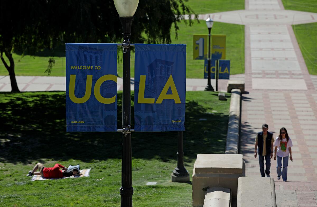 People lying on a lawn beyond a lamppost with 2 blue banners reading "UC" and "LA" in yellow, as 2 others pass on a walkway