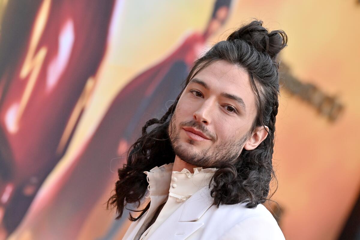 Ezra Miller gives a slight smile, wearing a white coat and high-collared shirt, their long hair worn down with a top knot