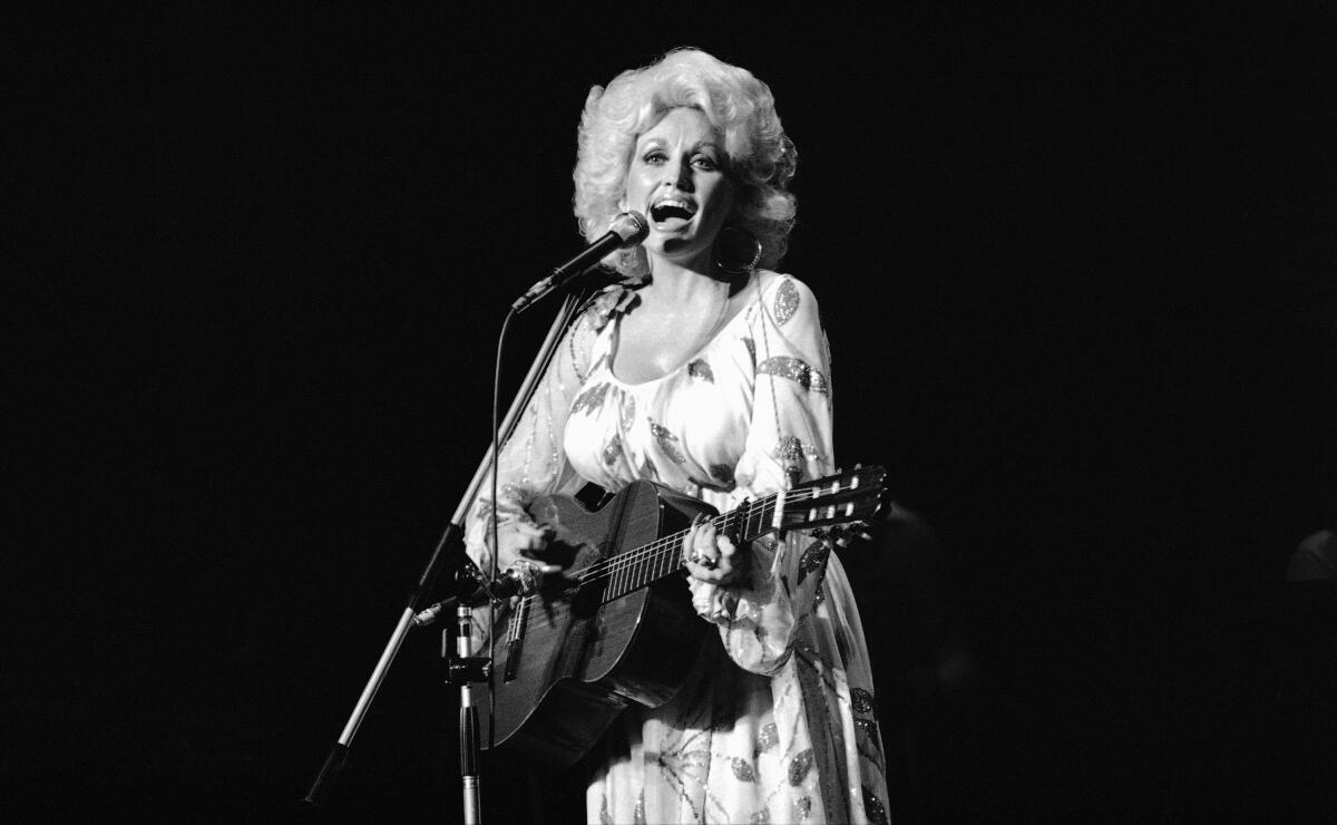 American singer Dolly Parton sings while playing a guitar and wearing a flowered dress.