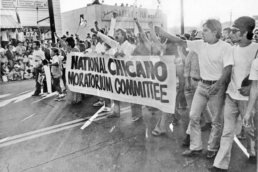 In a vintage black and white image, protestors carry a sign that reads "National Chicano Moratorium Committee"