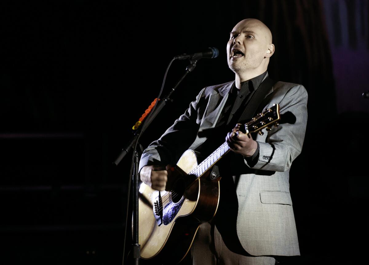 A man in a gray suit playing an acoustic guitar and singing into a microphone on a stage
