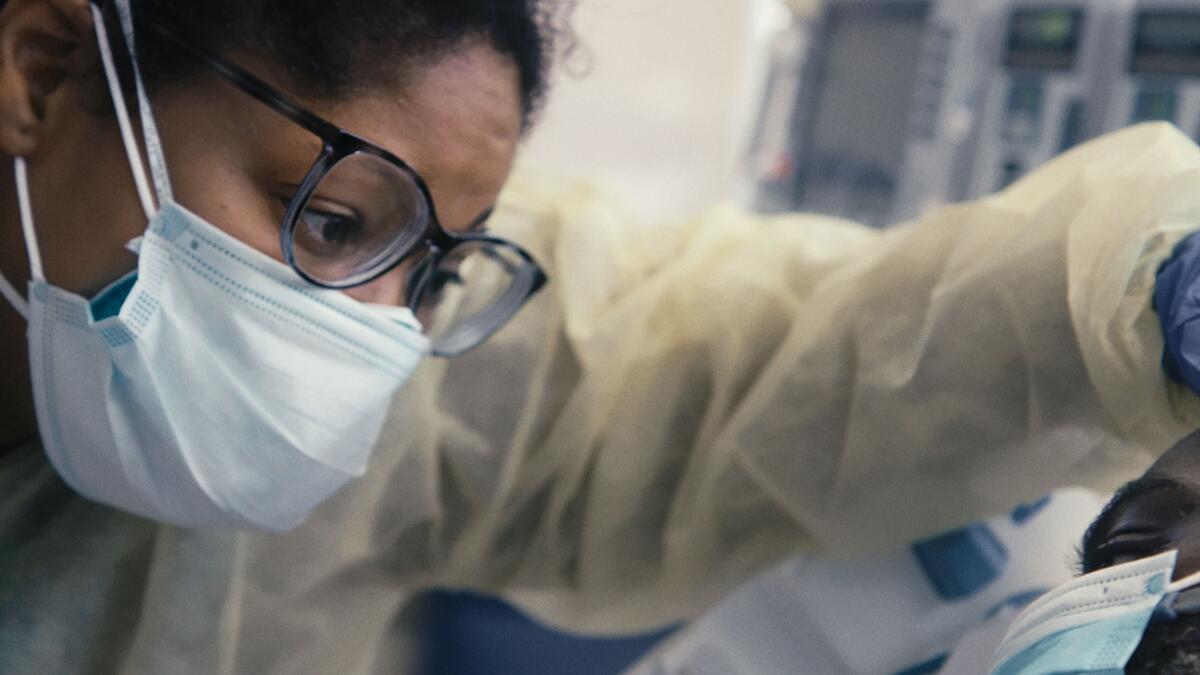 A person wearing glasses, a mask and protective attire stands over a patient in a hospital bed.