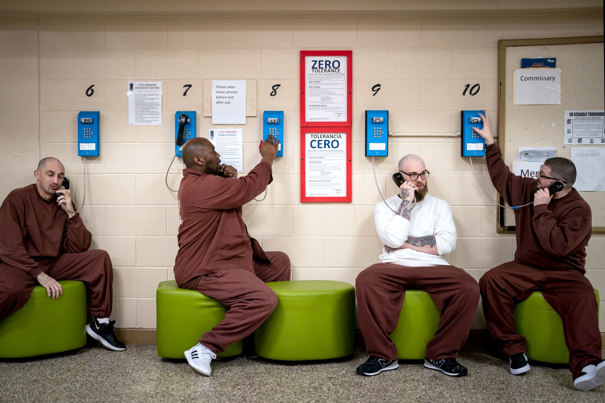 갈색 제복을 입은 4명의 수감자가 받침대에 앉아 그들 뒤에 있는 벽에 설치된 전화기로 각각 통화합니다.