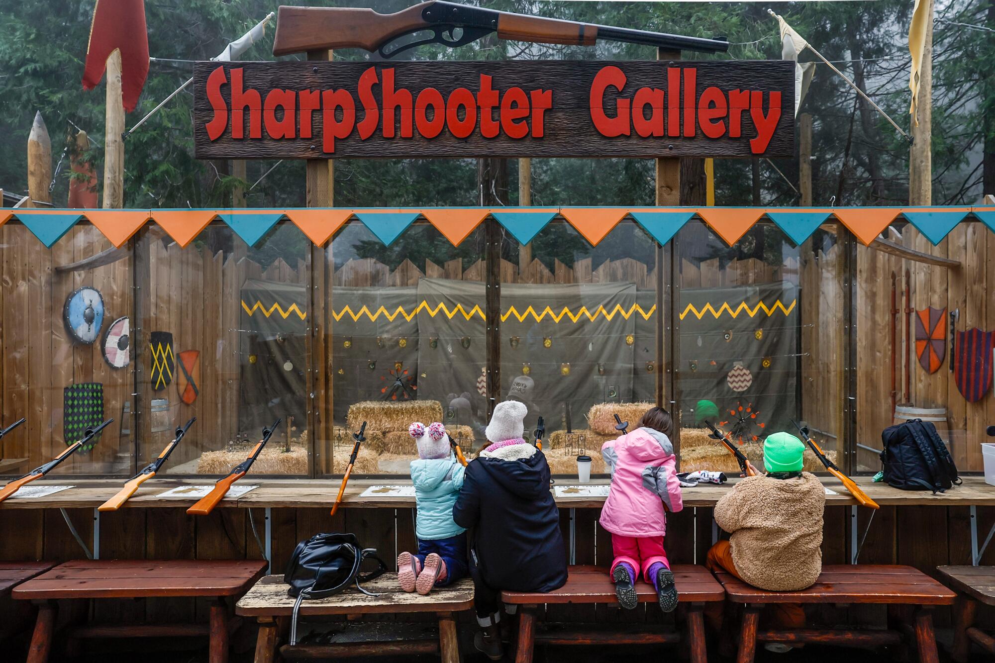Four children with their backs to the camera, at a play shooting range with a sign above them reading "Sharpshooter Gallery"