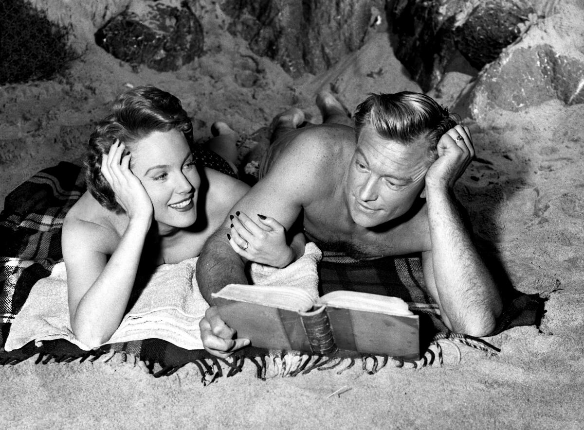 A woman and a man lie prone on the beach, their elbows propping up their heads; he holds a book.