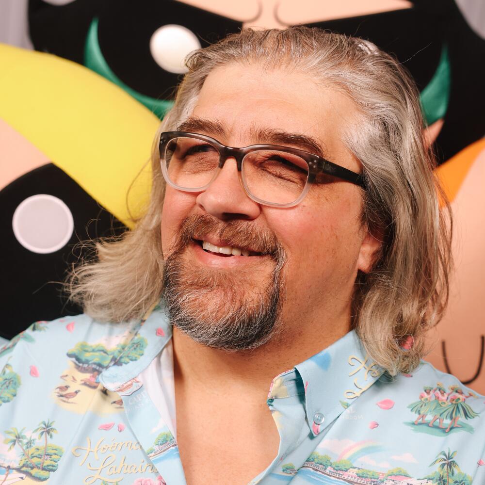 Craig McCracken in a blue Hawaiian shirt smiles and leans against giant plushes of Bubbles, Buttercup and Blossom.