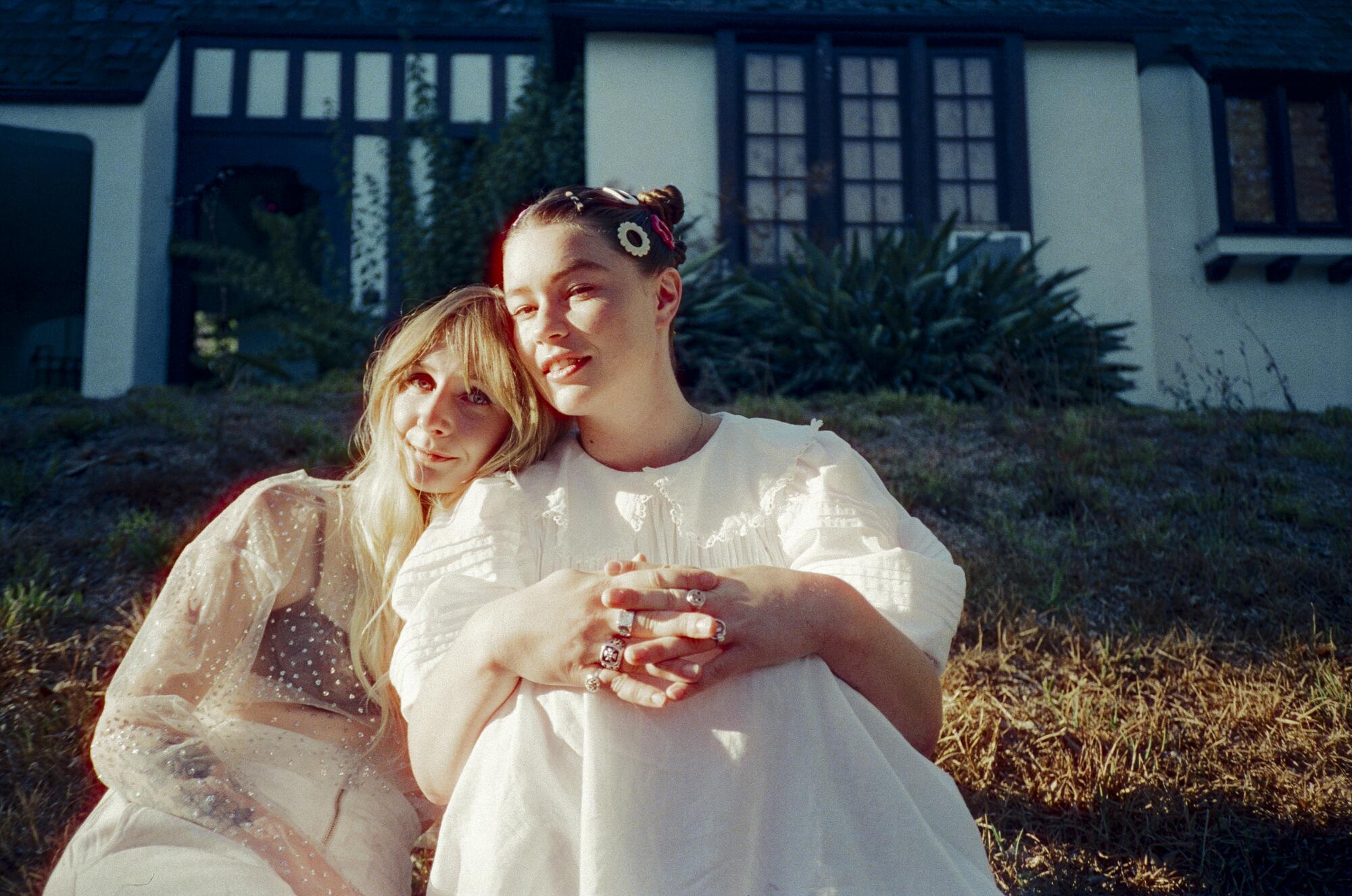 Two women sit side by side outside, one laying her head on the other's shoulder