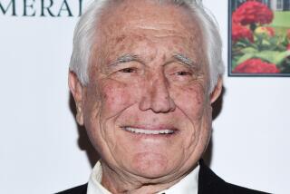 George Lazenby smiles and poses as he looks off camera while wearing a black and white tuxedo