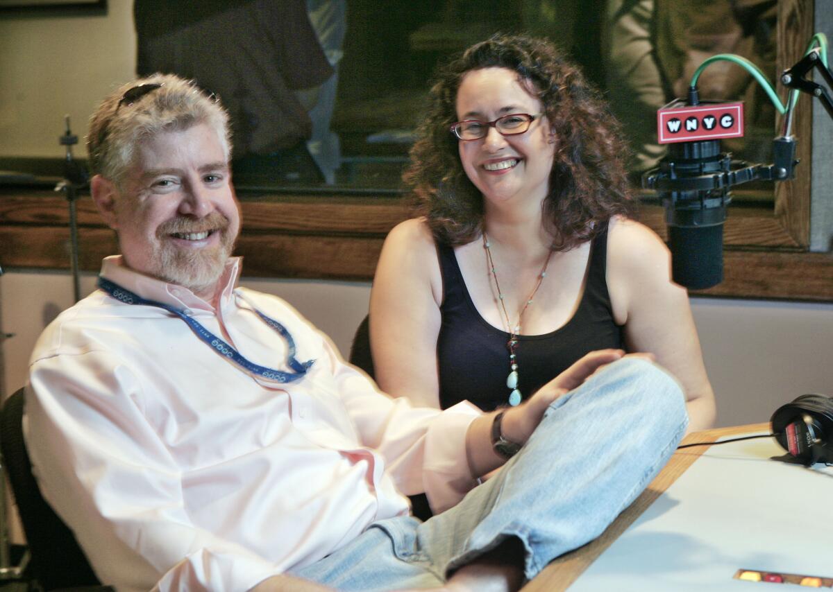 A man and a woman pose, seated, behind a microphone that says "WNYC."