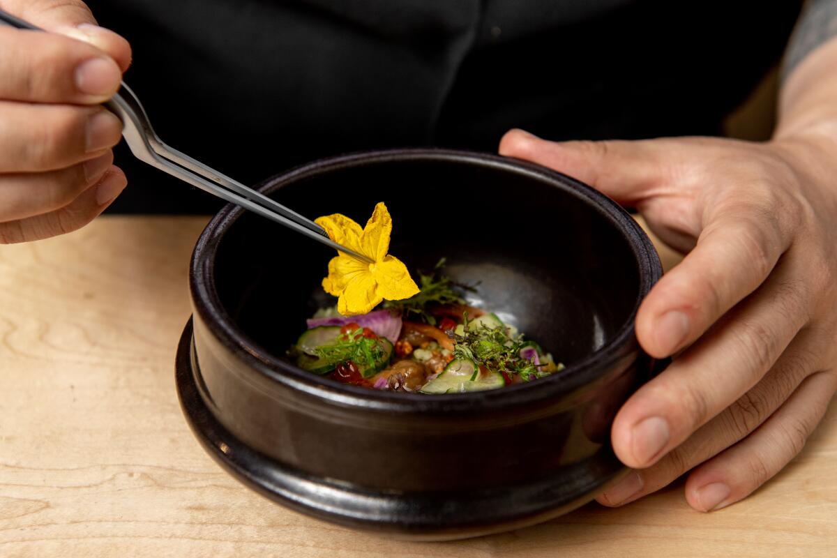 Closeup photo of a chef's hand using tweezers to place a yellow flower in a black bowl containing bibimbap