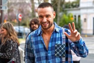 Liam Payne with a shaved head wearing a blue flannel shirt smiling and posing with his right hand up