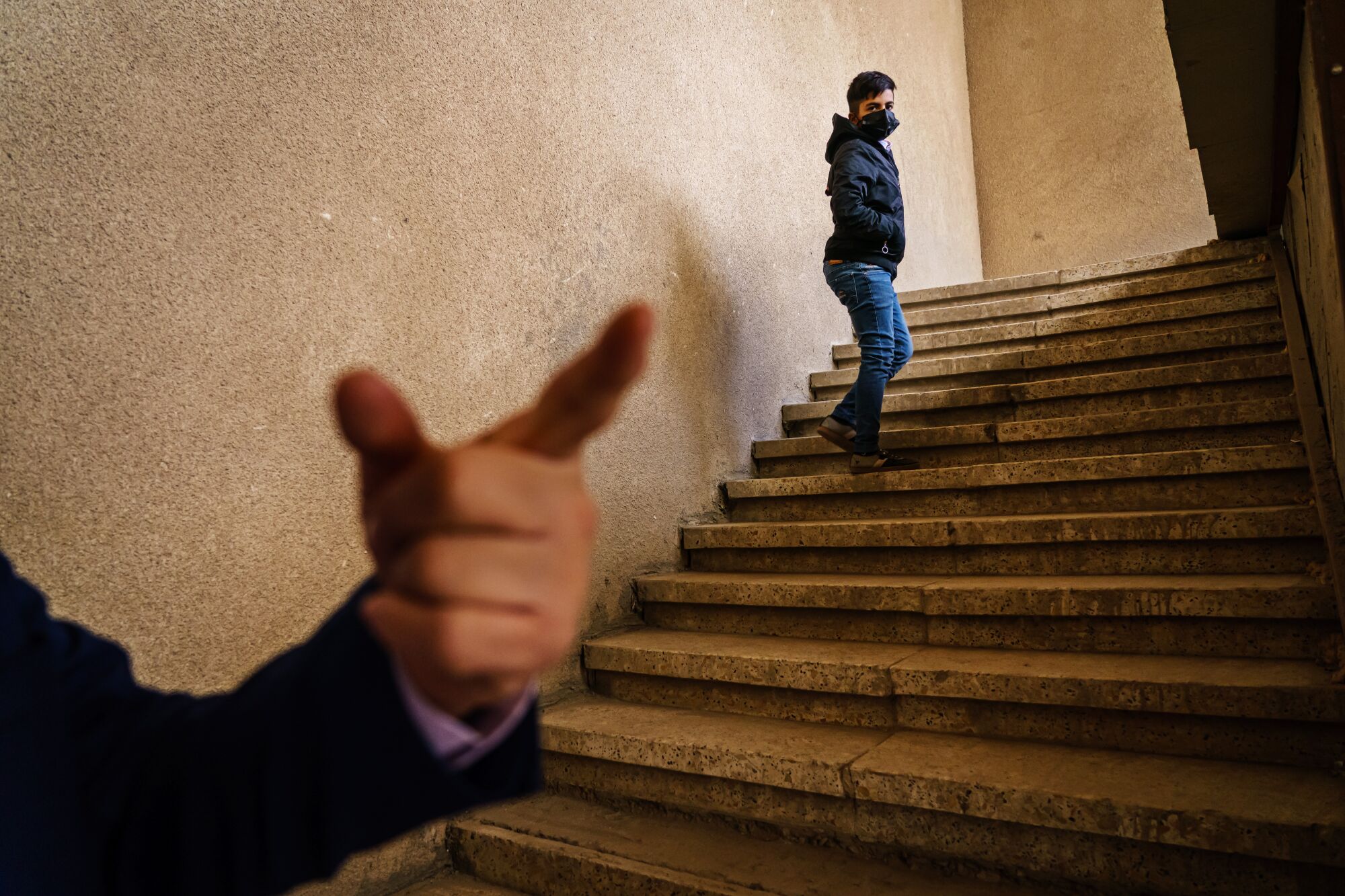 A masked person stands on a flight of steps looking toward an unseen person gesturing with one hand 