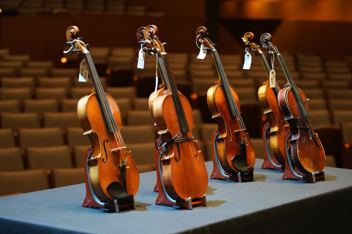 Six tagged violins sit on stands on a stage.