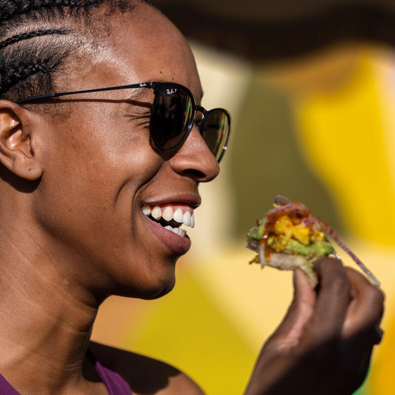 A smiling woman in sunglasses enjoys a slice of avocado toast.