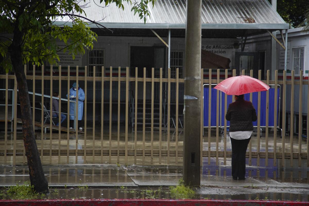 En el Hospital General de Tijuana, una mujer con un paraguas rojo observa mientras un paciente es examinado por un trabajador de la salud del hospital.