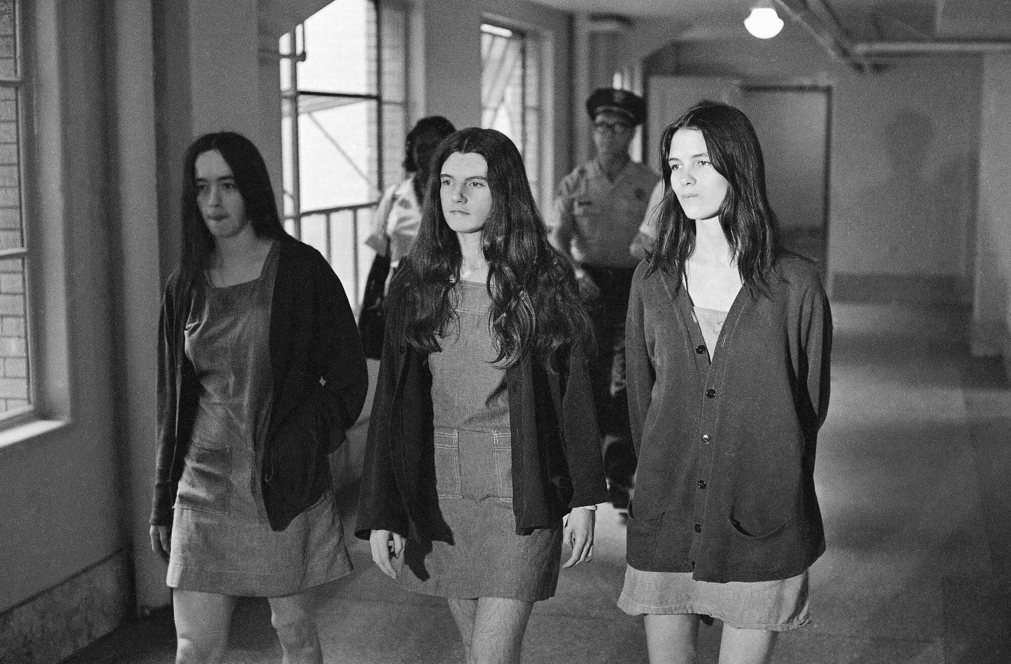 Three women walk down a hallway, with two guards behind them.