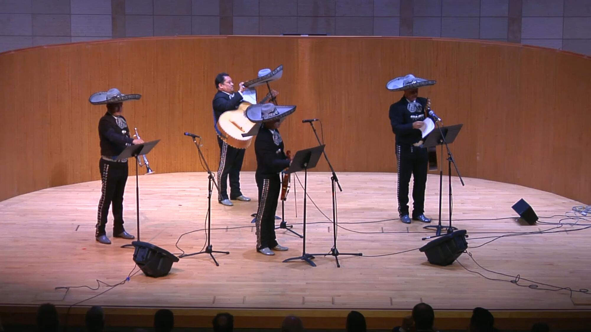 A still from a video shows four performers from a mariachi troupe on stage, one of whom is adjusting his sombrero.
