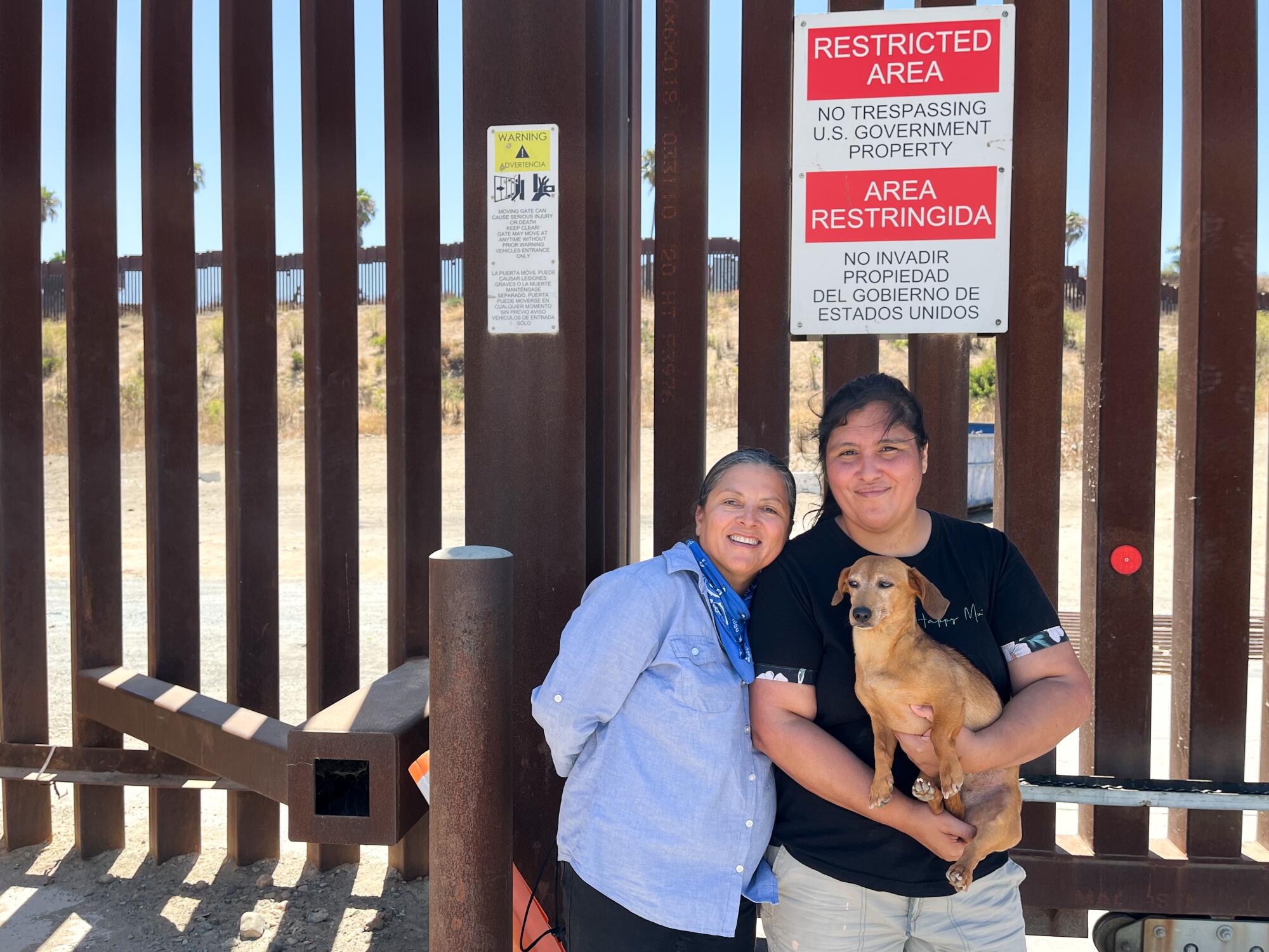 A smiling woman in a blue shirt, left, and a woman in a dark top, holding a dog, stand in front of a slatted tall fence