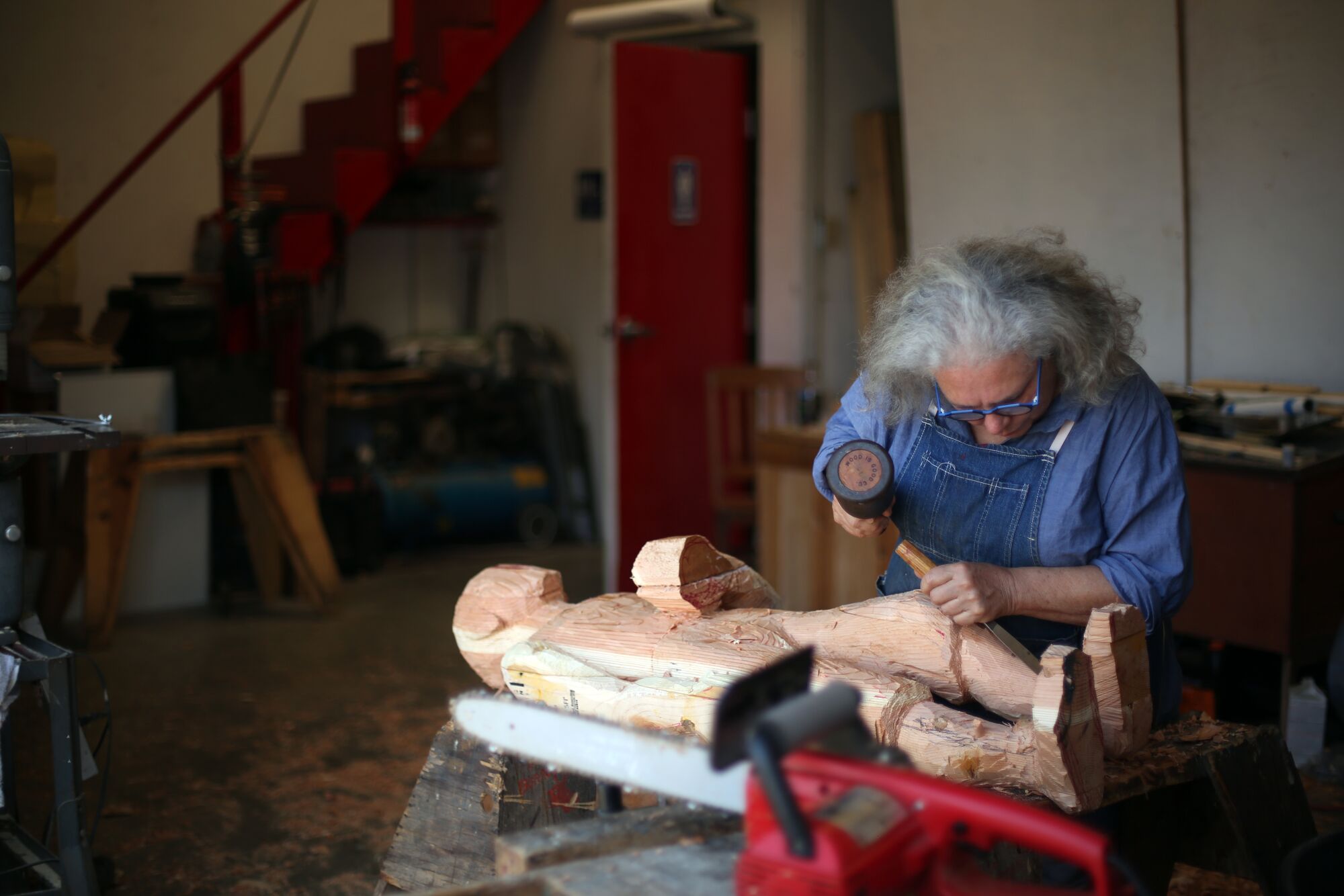 Alison Saar, in overalls, chisels at a wooden sculpture in a warehouse studio