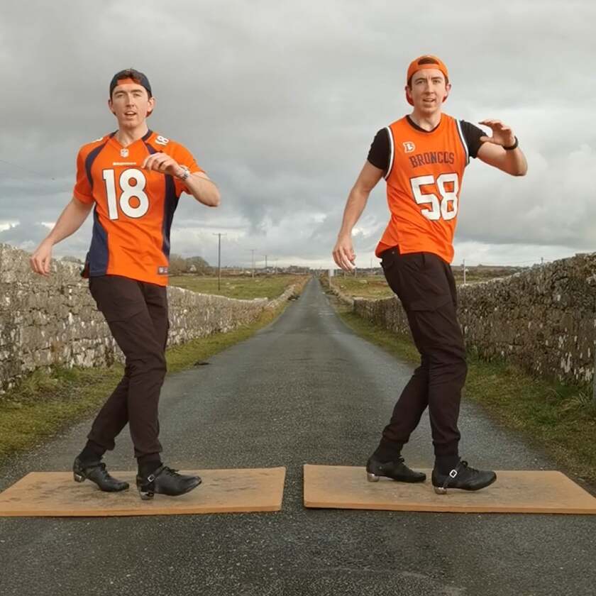 Two men in football jerseys and tap shoes dance on boards in the middle of an empty road bordered by low rock walls.
