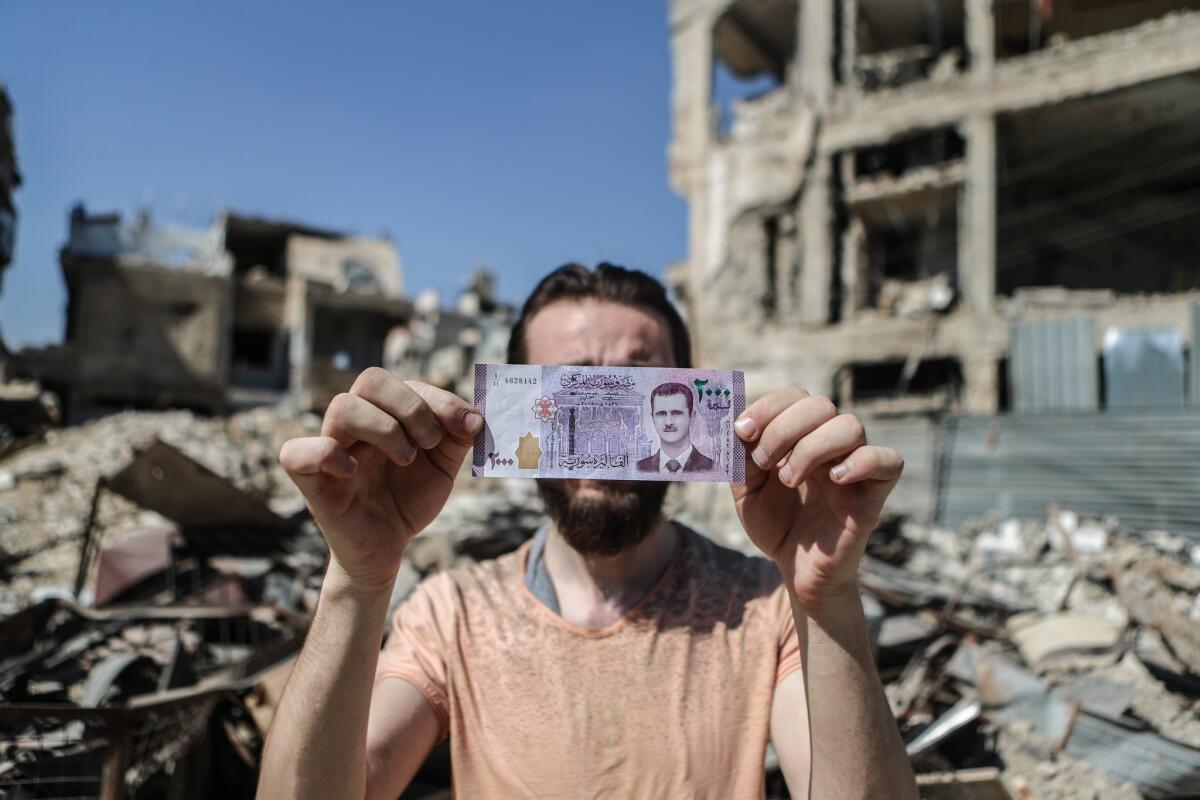 A Syrian man holds up a 2,000-pound banknote, featuring President Bashar Assad, in front of damaged buildings, in Duma, Syria, in 2017.