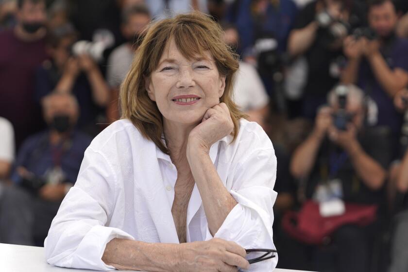 Jane Birkin smiles in a white shirt while resting her chin on her hand in front of a crowd of photographers.