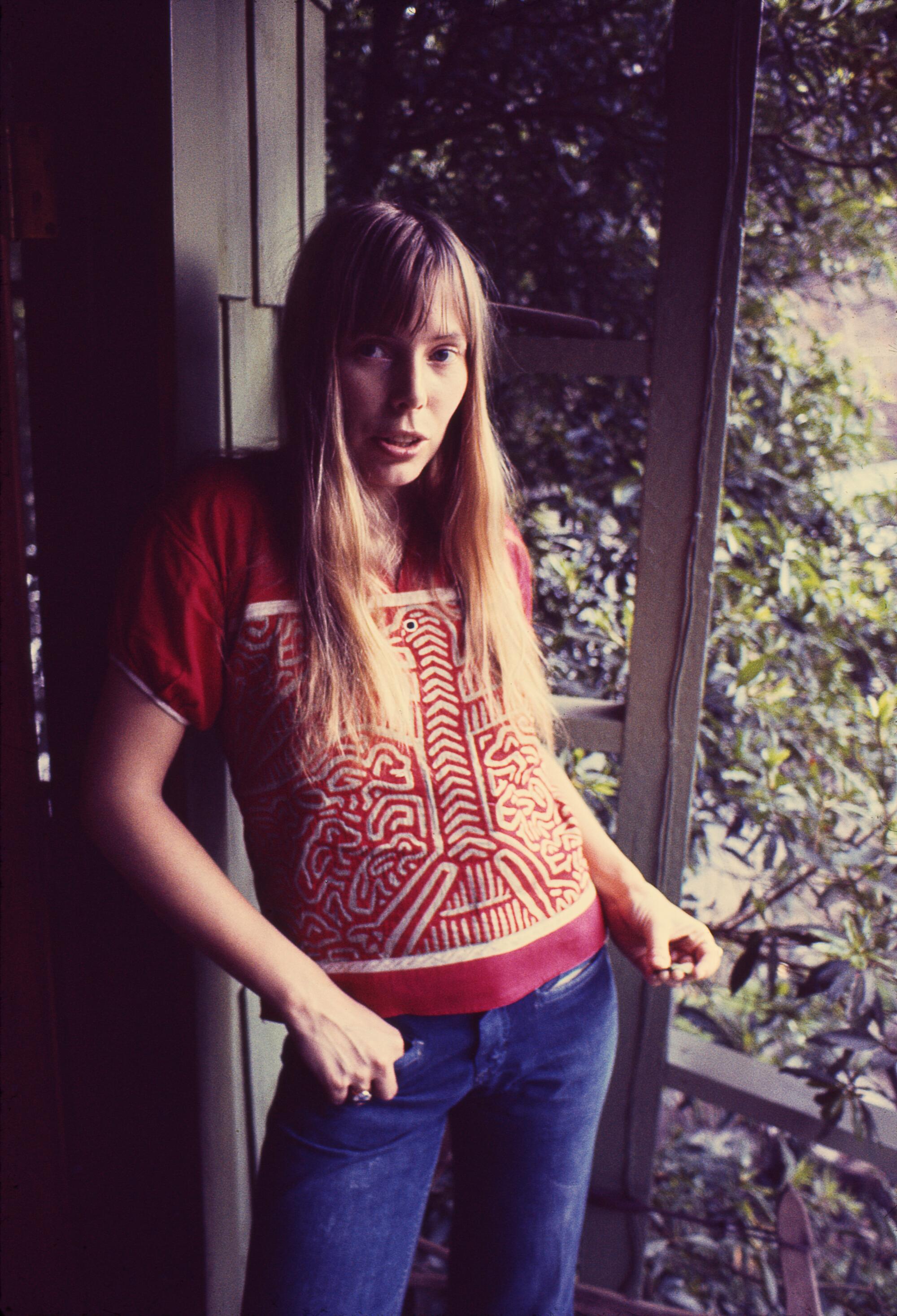 A young Joni Mitchell wearing jeans, standing against a brown shingle house.  
