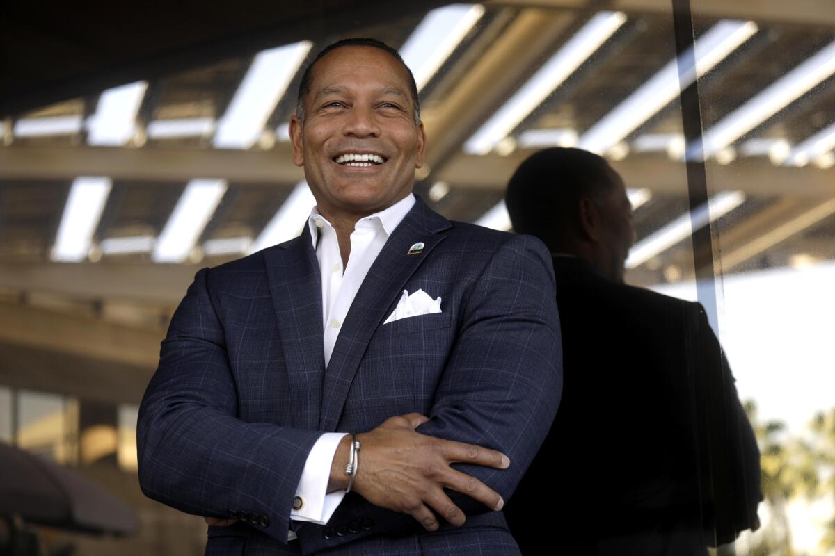 A man wearing a dark jacket and white shirt smiles while standing. 