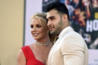 Britney Spears is smiling and wearing a red dress while posing next to Sam Asghari who is in a cream suit