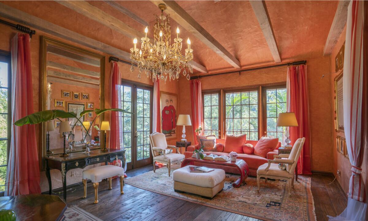 A living room in shades of orange with a chandelier, furniture and windows overlooking foliage.