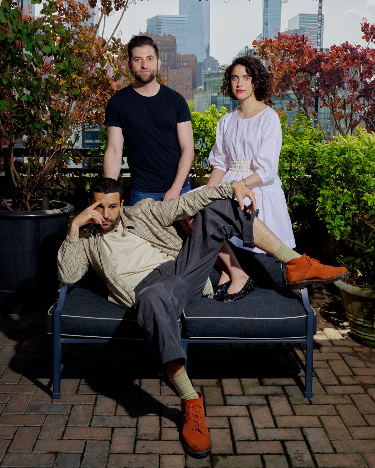 A man and a woman stand behind a man reclining on an outdoor couch, with the New York City skyline in the background.