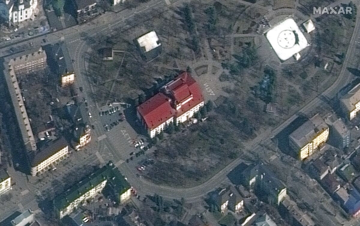 A bird's-eye view of a red-roofed building, flanked by letters in white on the ground, and buildings nearby