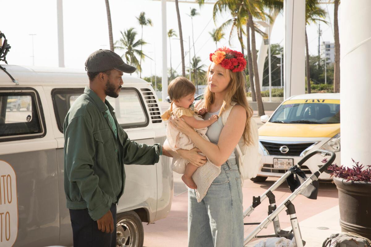A man in a baseball cap and a woman holding a baby stand next to a van outside a building.