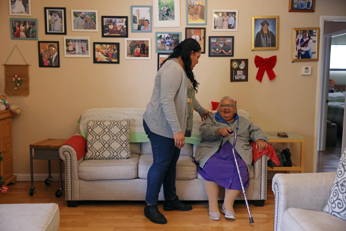 Helen Cordova stands next to her mom, who is seated in a living room.