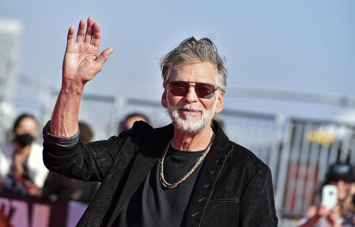 A man with gray hair and a beard waves as he walks.