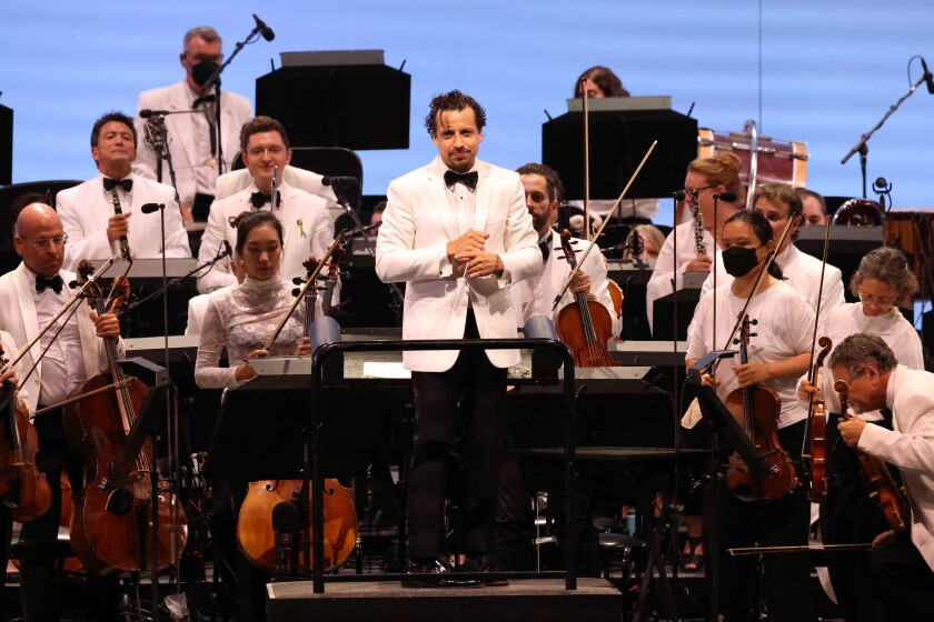 A man in a white jacket and black bow tie at the conductors podium with similarly attired musicians behind him.